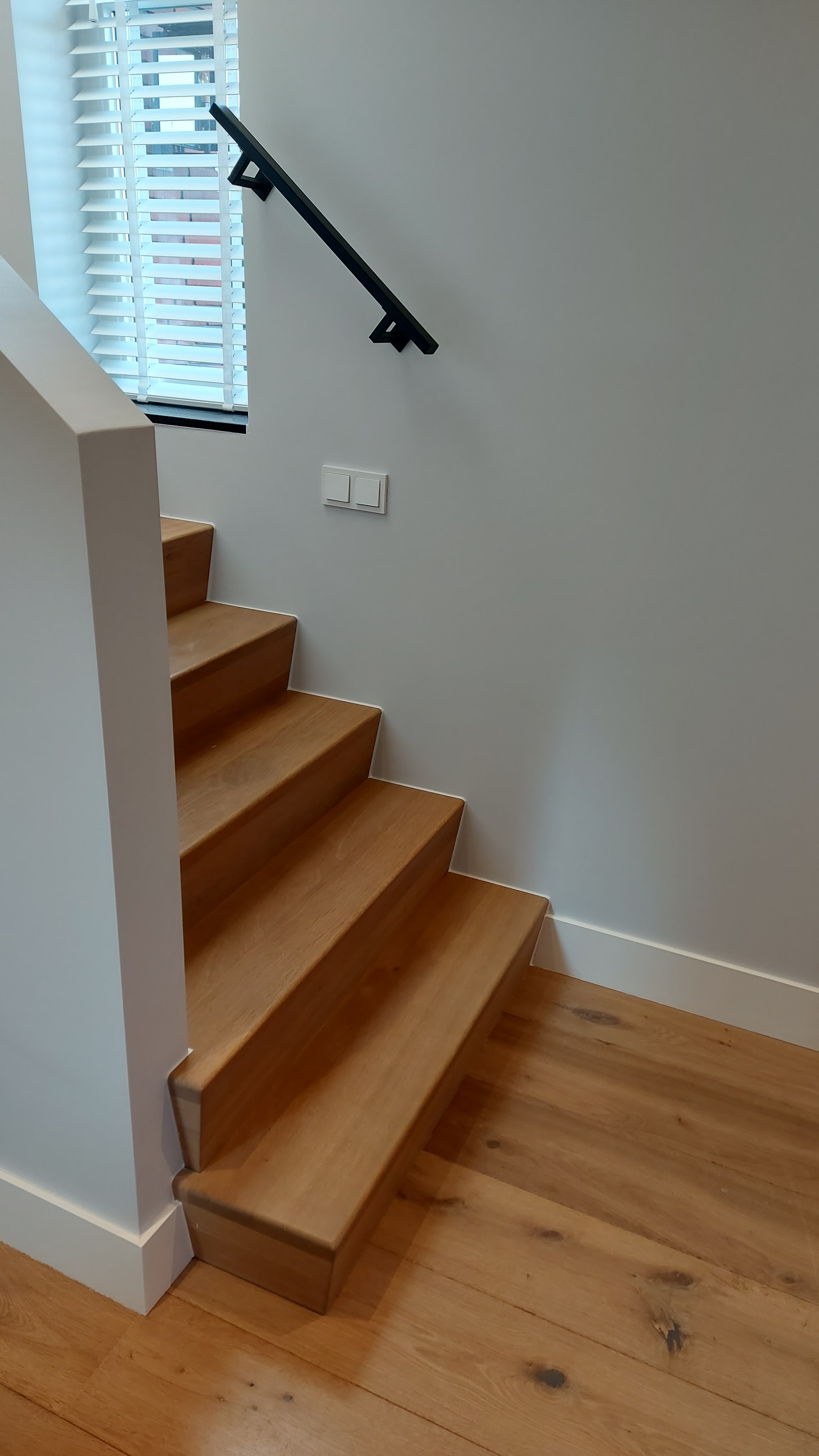 Eiken trap langs een grijsgroene wand met witte plinten en een zwarte leuning